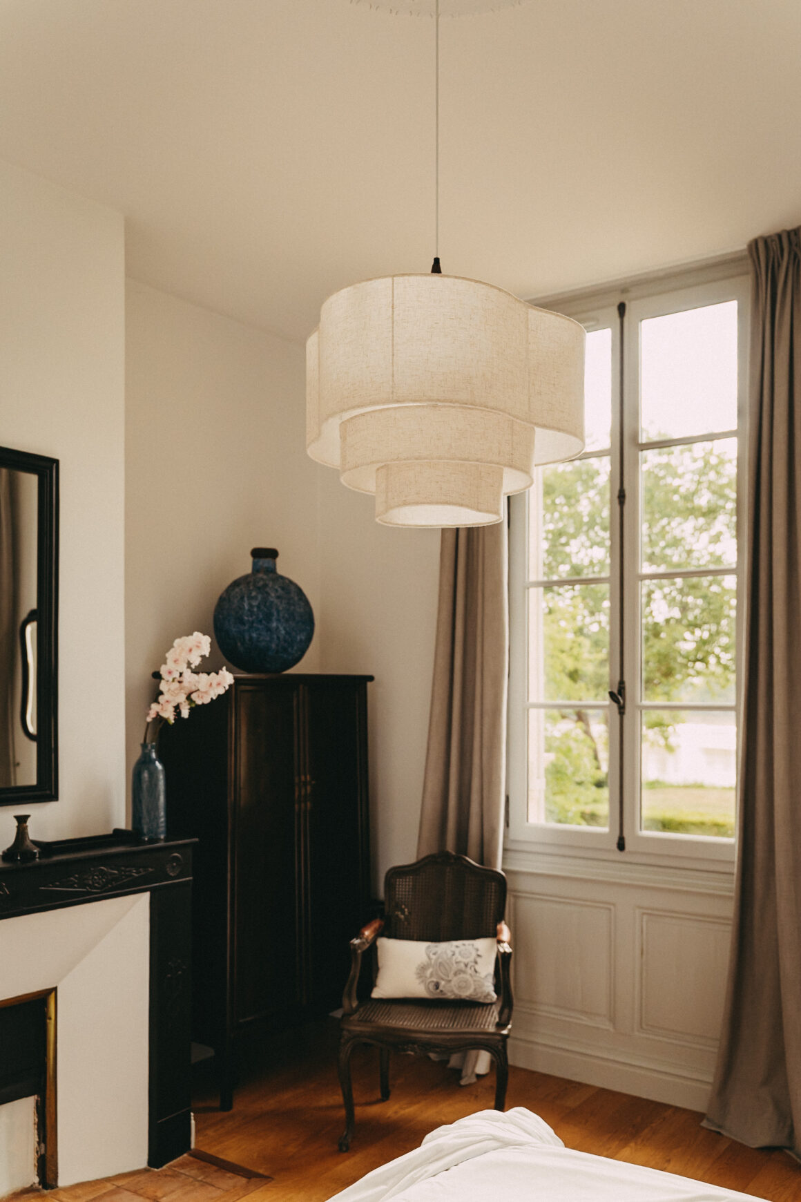 Chambre lumineuse du Château Larteau avec grande suspension en tissu, fauteuil ancien et mobilier en bois sombre.