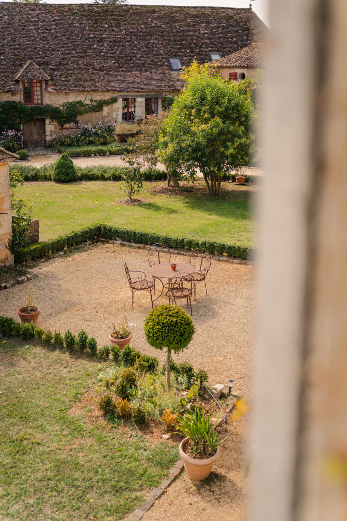 Vue sur la cour intérieure du Domaine de Lavernelle, avec une table et des chaises en fer forgé, un jardin arboré et des bâtiments en pierre.