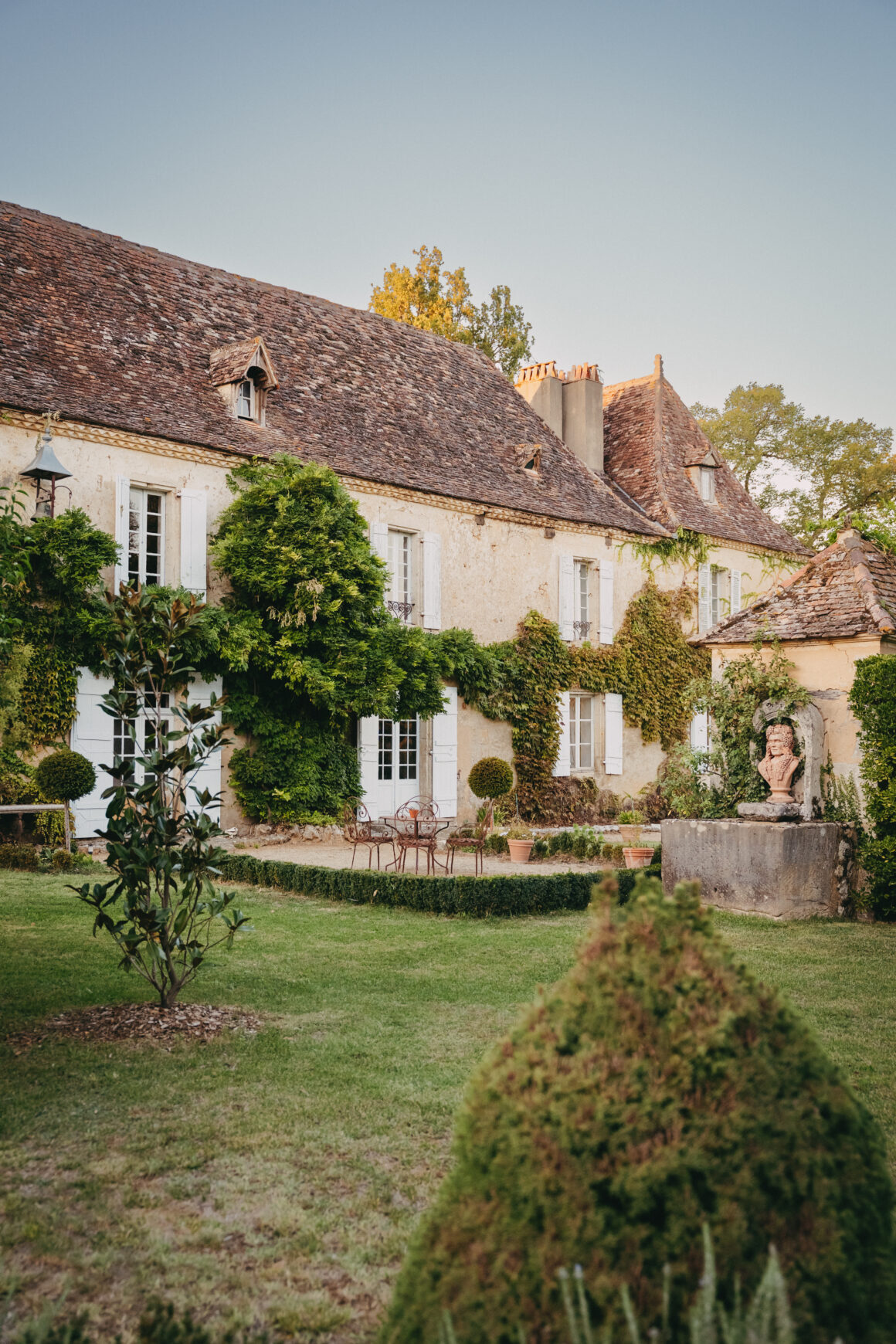 Façade en pierre du Domaine de Lavernelle, avec ses volets blancs, sa végétation grimpante et son jardin paysager.