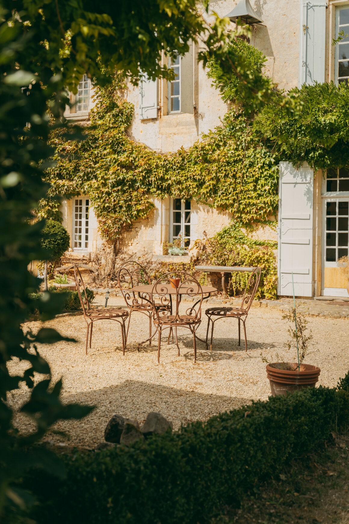 Table et chaises en fer forgé dans la cour ensoleillée du Domaine de Lavernelle, devant une façade couverte de vigne vierge.