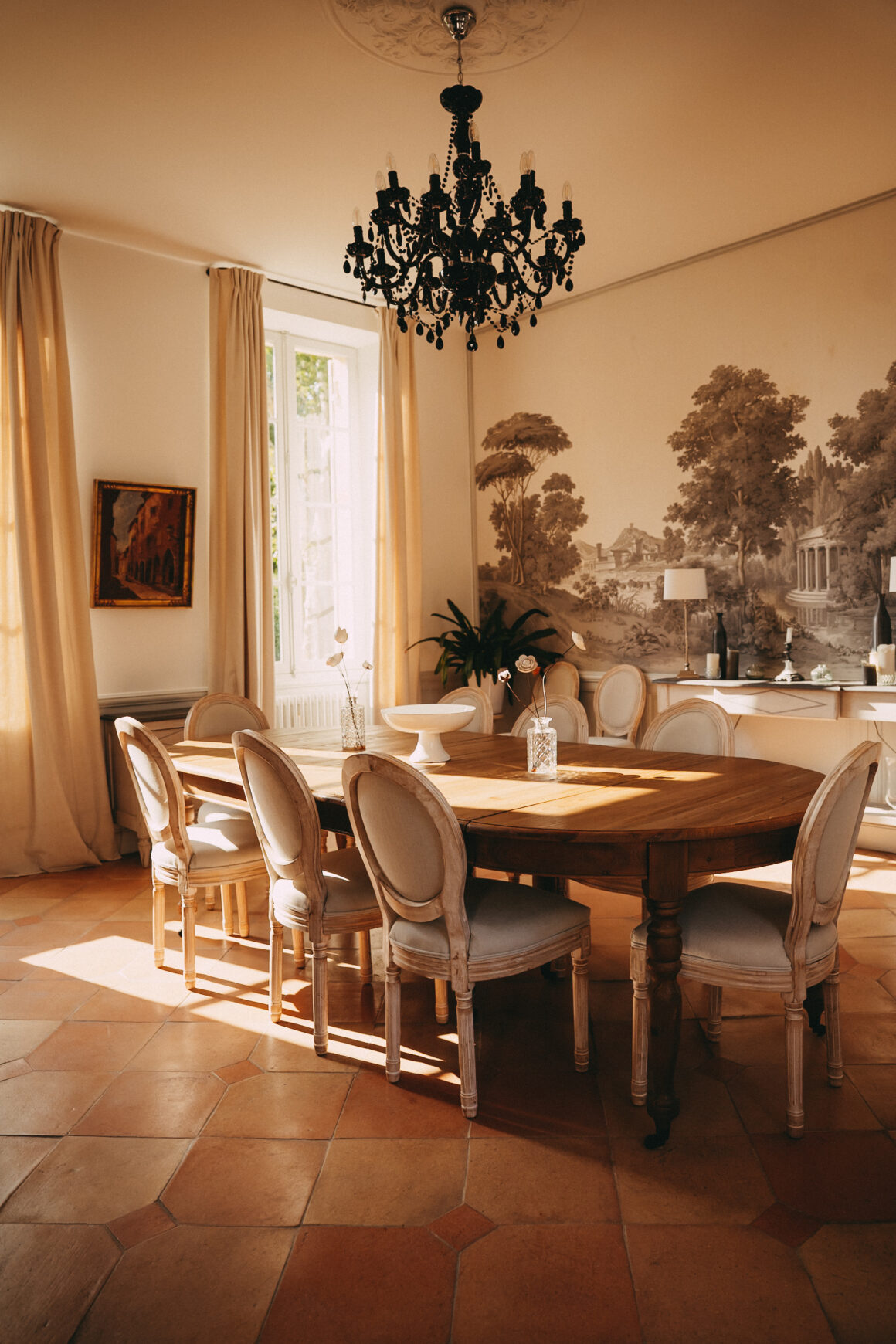 alle à manger du Château Larteau près de Bordeaux, baignée de lumière naturelle, avec table en bois, lustre noir et décor panoramique.