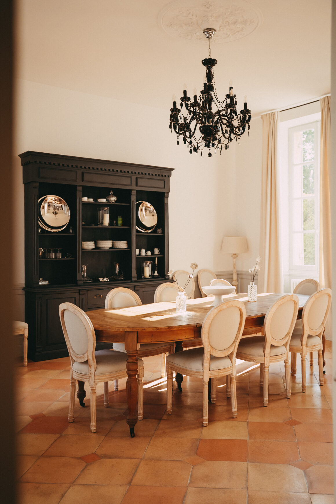 Salle à manger du Château Larteau avec grande table en bois, chaises médaillon, vaisselier ancien et lustre noir.