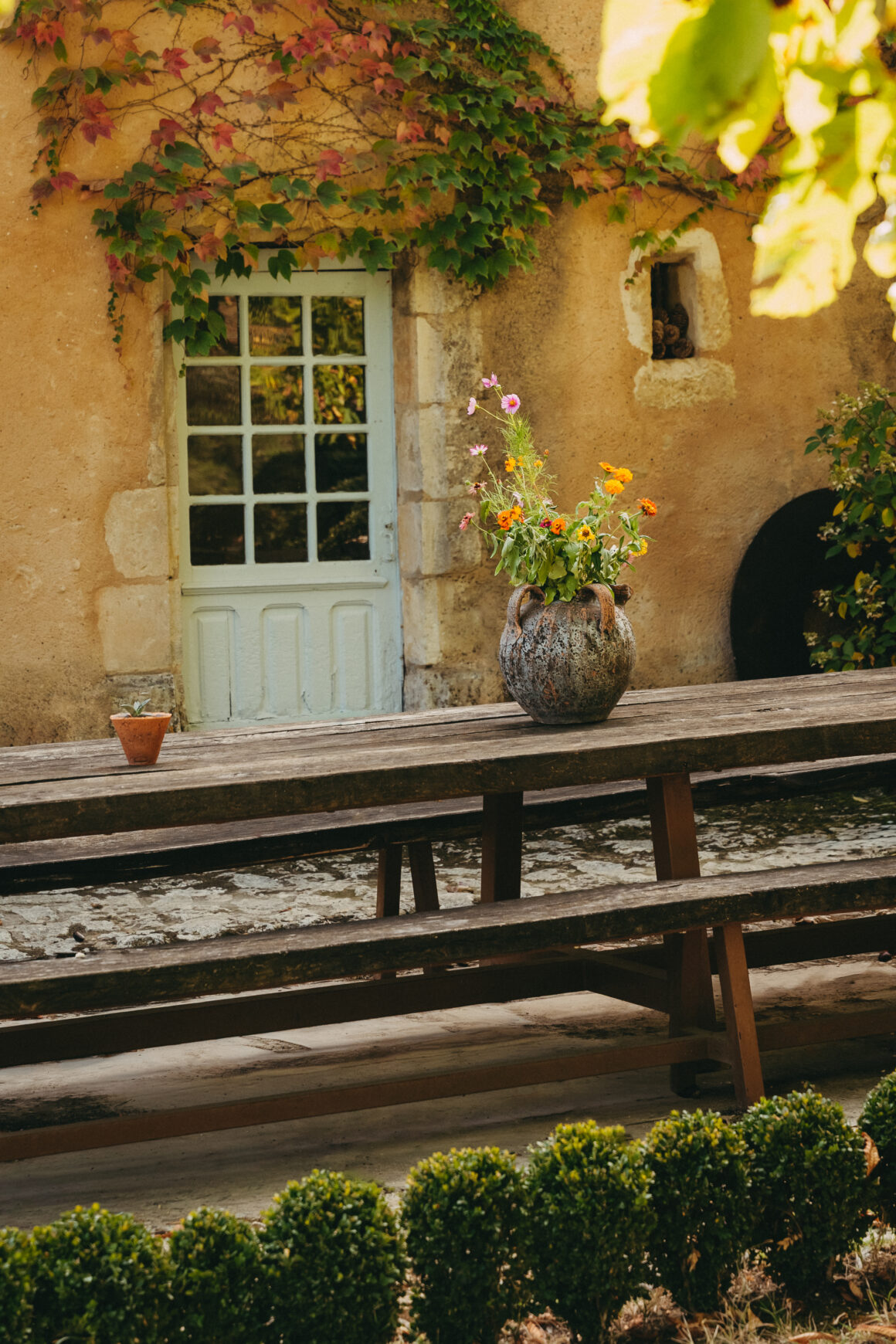 Grande table en bois ornée d’un vase de fleurs, devant une façade ancienne aux volets clairs du Domaine de Lavernelle.